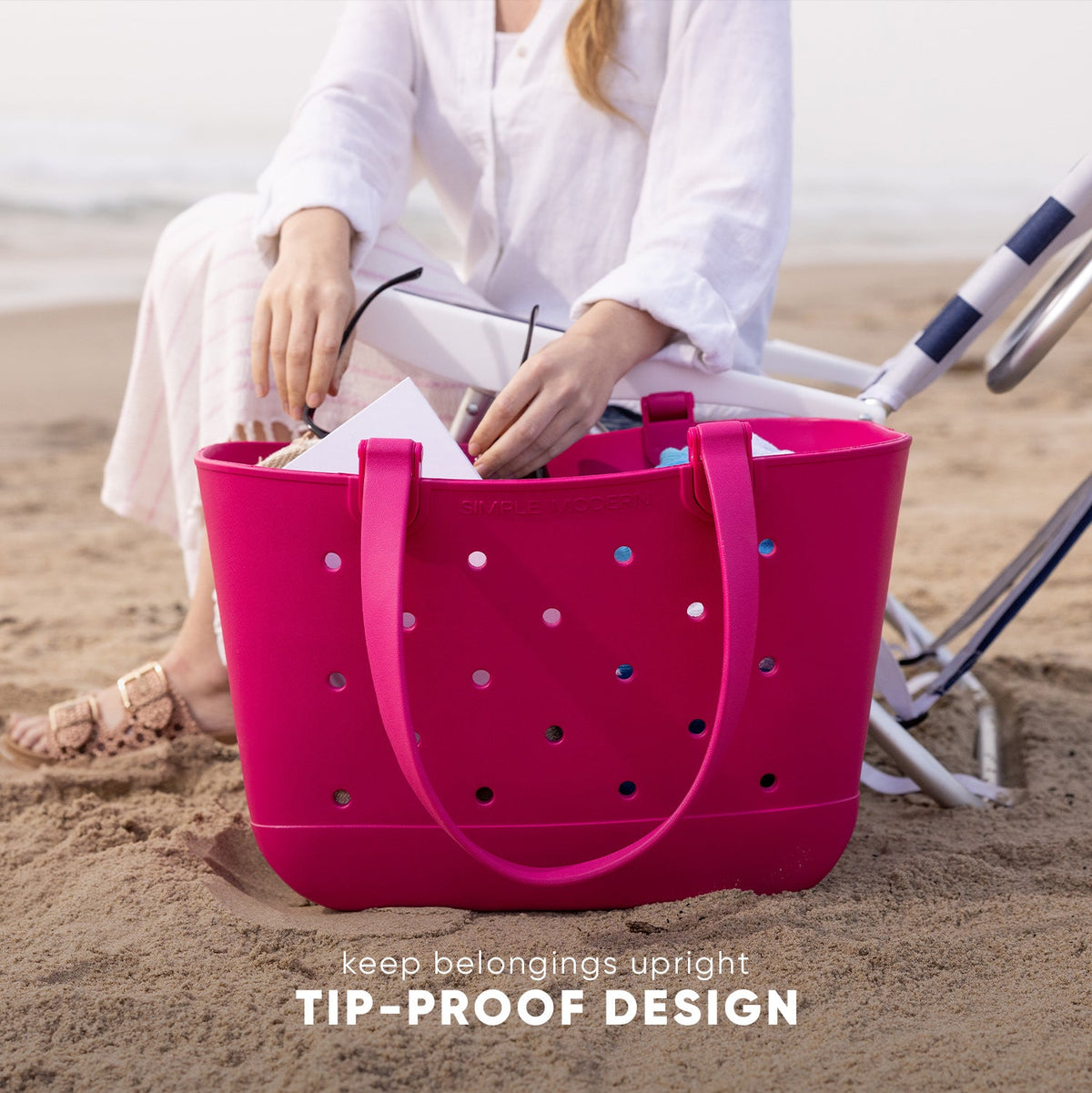 Bright hot pink Simple Modern Getaway Bag with tip-proof design held open on a beach, revealing white items inside. Navy striped beach chair visible in background. Text reads "keep belongings upright TIP-PROOF DESIGN."