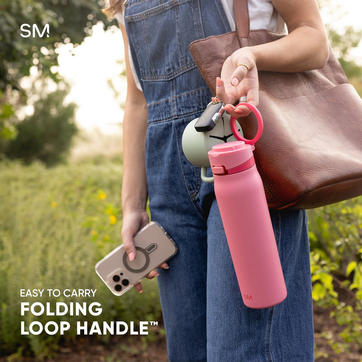 Person holding pink insulated water bottle with folding loop handle, smartphone, and yellow flowers in garden setting. Lifestyle product photo showcasing portable water bottle design.