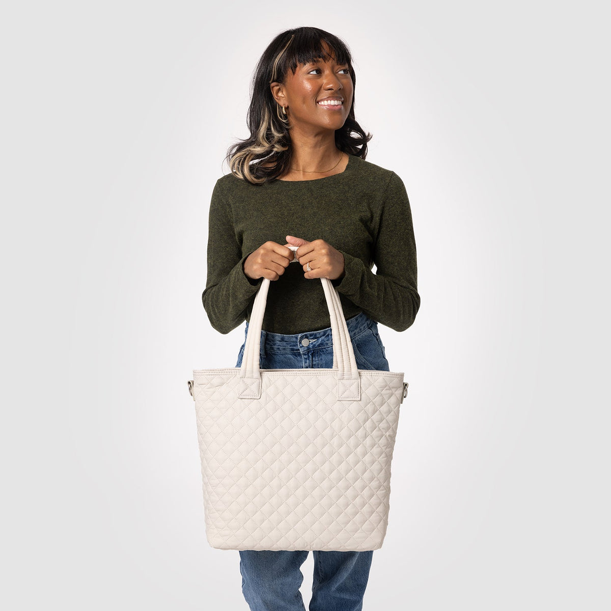 Cream quilted tote bag held by model wearing olive-green long-sleeved top and medium-wash blue jeans against neutral background
