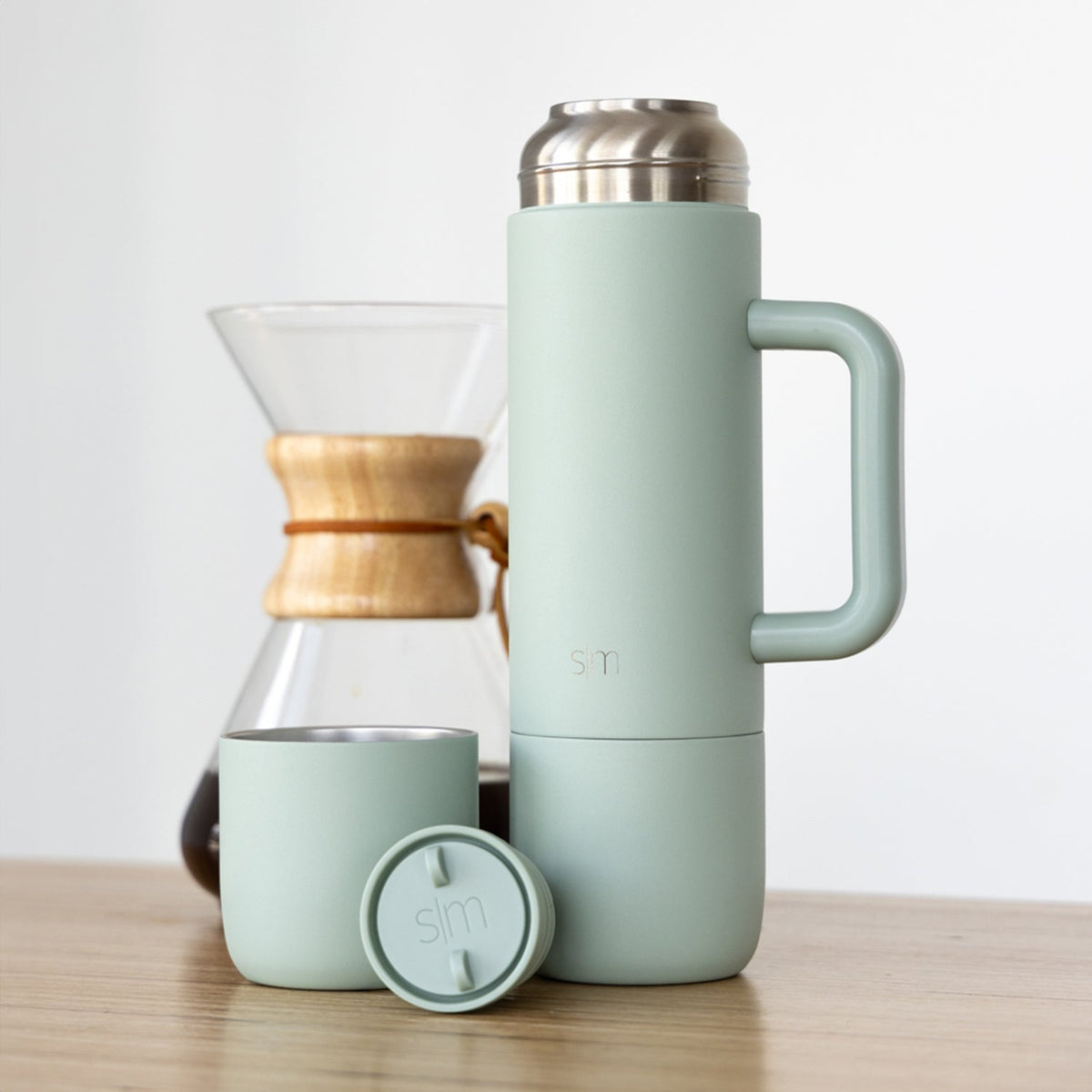 Sage green insulated thermos next to matching cup and Chemex coffee brewer with wood-tone band, on white background