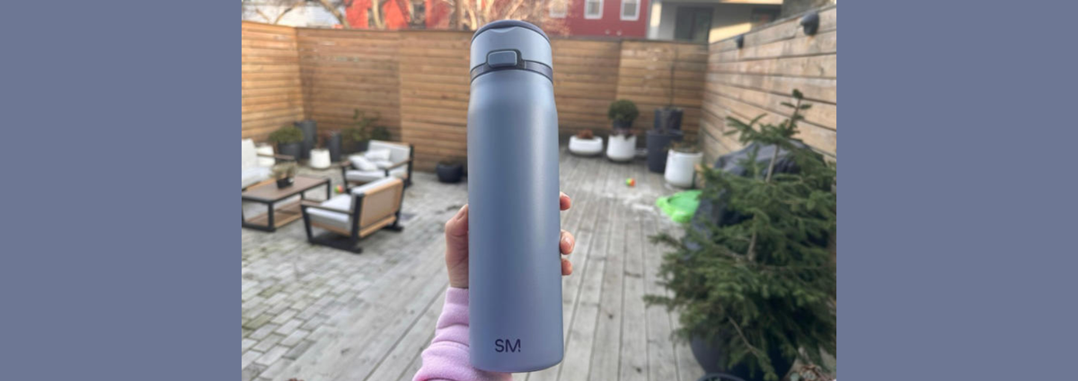 Person holding light blue Simple Modern insulated water bottle with flip-top lid on residential patio with wooden deck, fence, and potted plants in background.